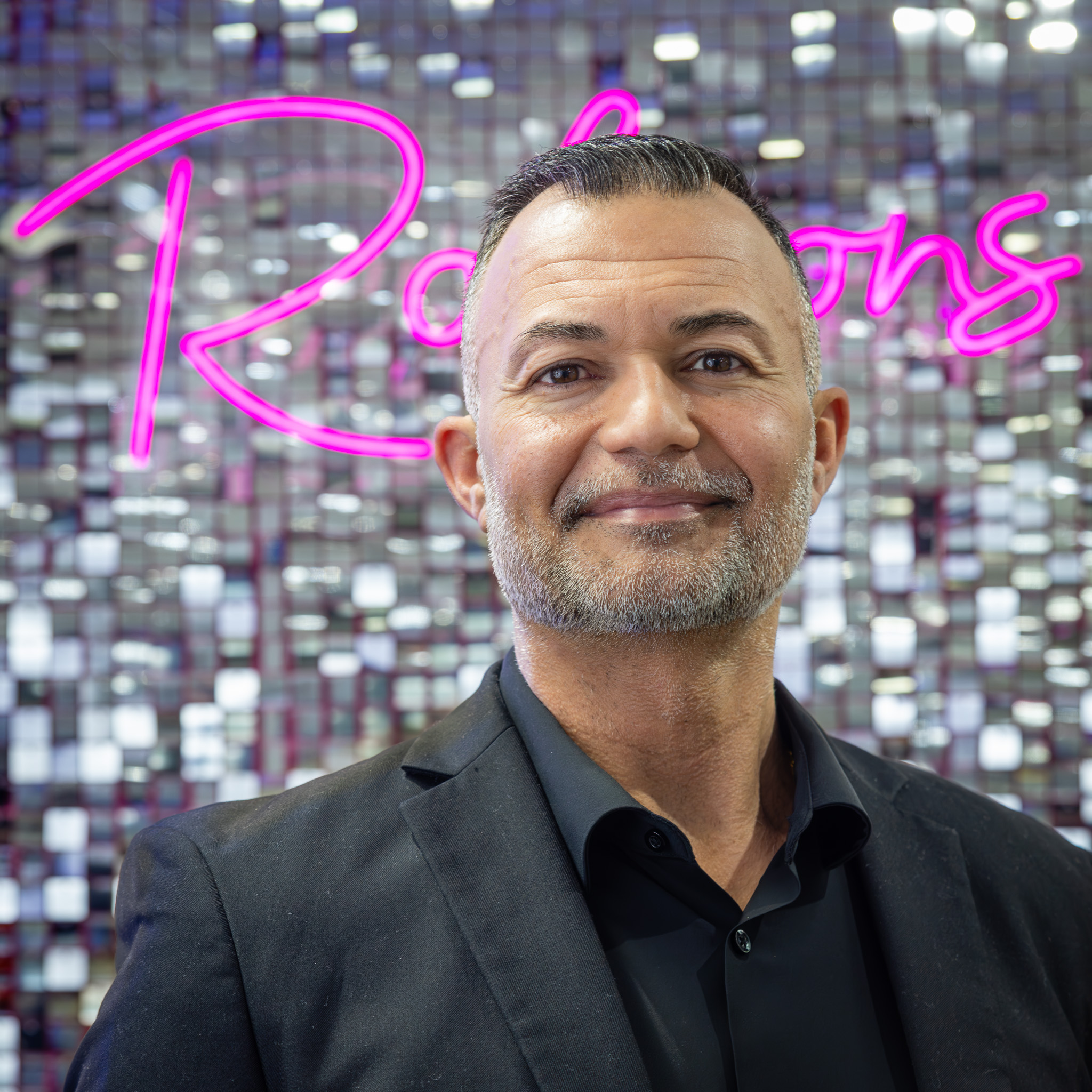 Man in a black suit standing before reflective square tiles with a pink neon “Robsons” sign glowing behind him.