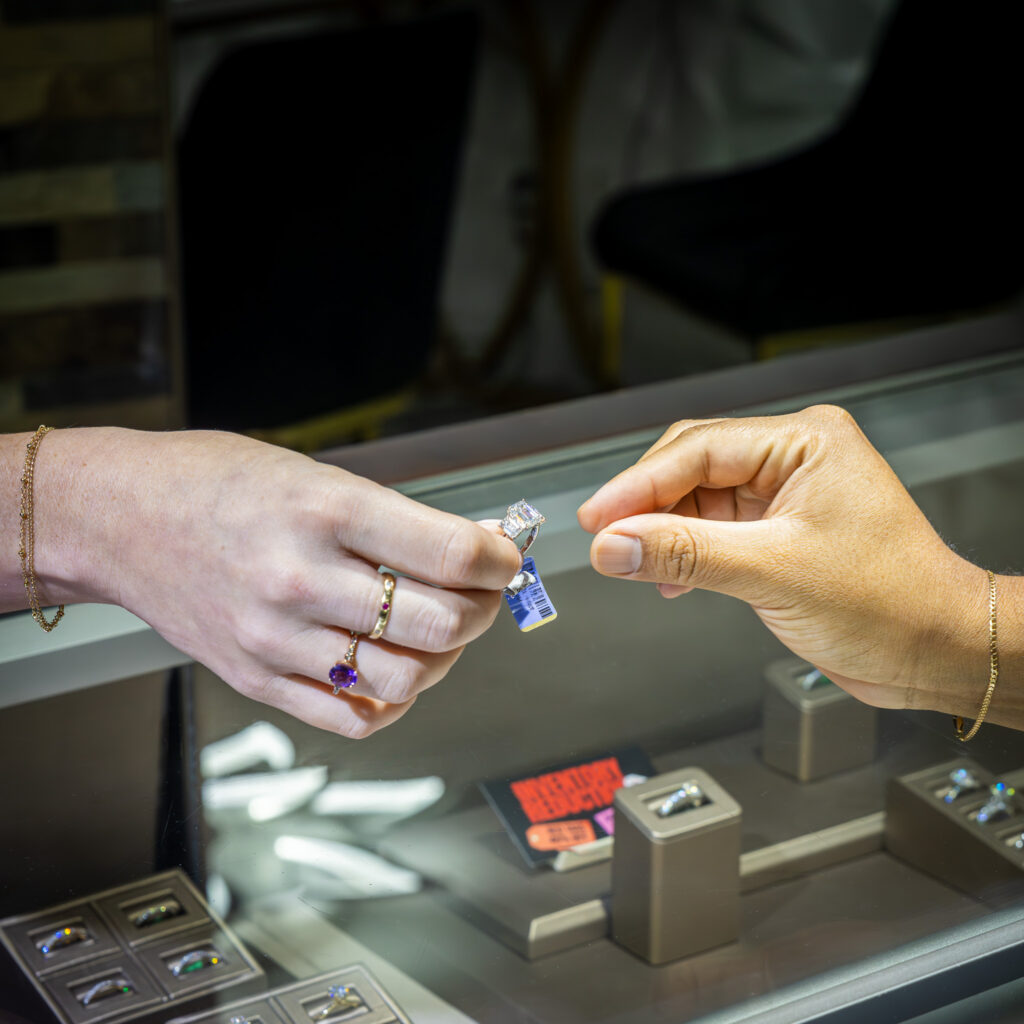 Hands exchanging a diamond ring over a glass jewelry counter with multiple rings on display in a retail store.