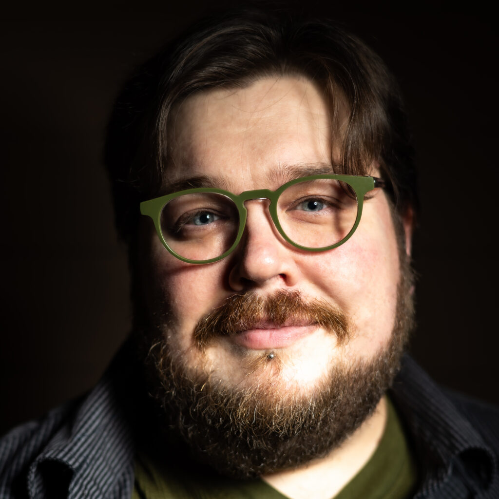 Close-up portrait of a bearded person wearing green-framed glasses, lit dramatically against a dark background.