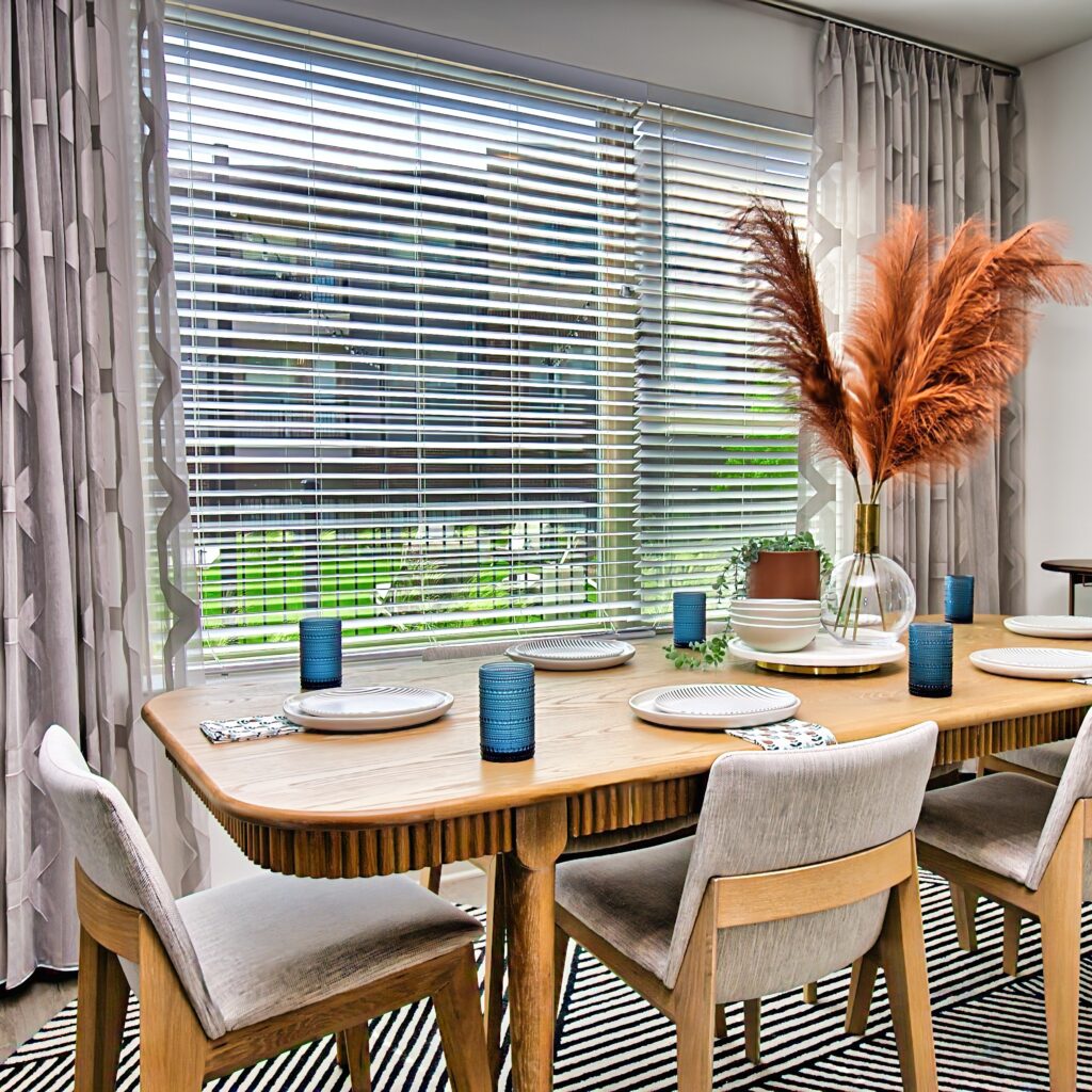 Modern dining room with a wooden table set for six, featuring blue glasses, patterned napkins, and a pampas grass centerpiece beside a large window.