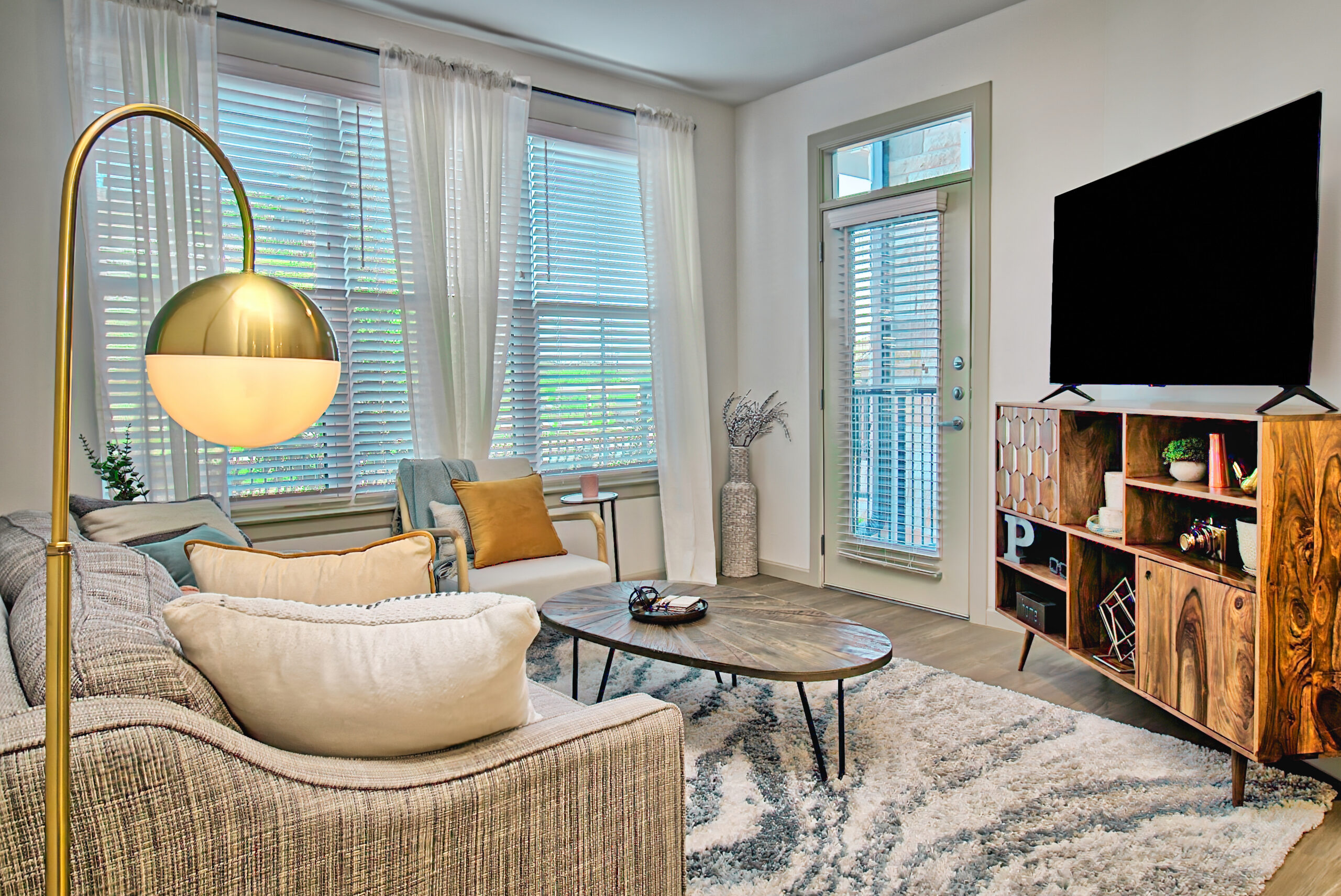 Modern living room with a gray sectional sofa, blue accent chair, gold floor lamp, and a wooden media console beneath a wall‑mounted TV, lit by large windows with sheer curtains.