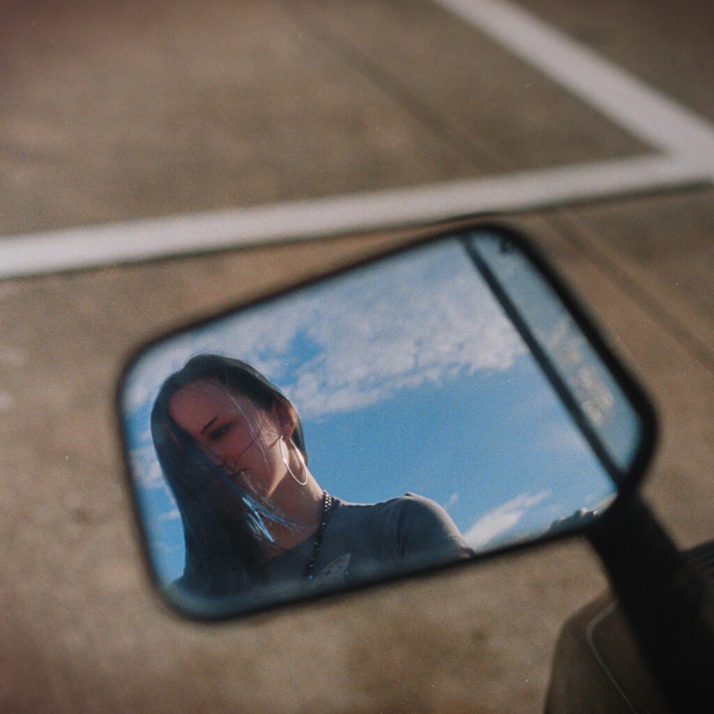Reflection of a person in a motorcycle side mirror against a bright sky in a parking lot.