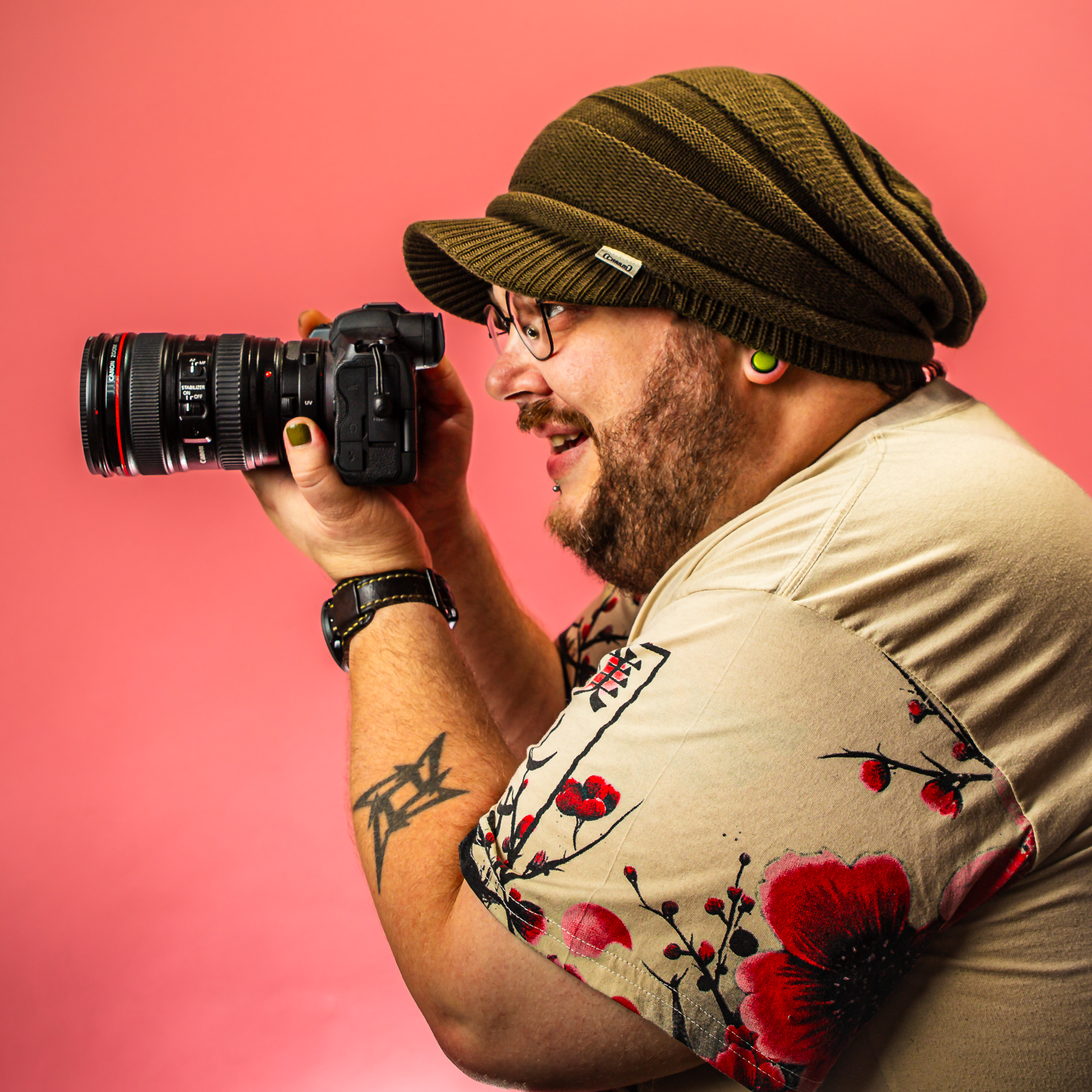 Person holding a DSLR camera against a pink background, wearing a green beanie, glasses, and a floral beige shirt while photographing.